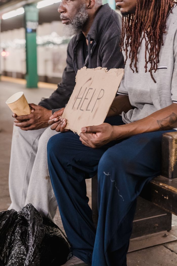 Two homeless individuals sitting on a bench holding a help sign in a city setting.